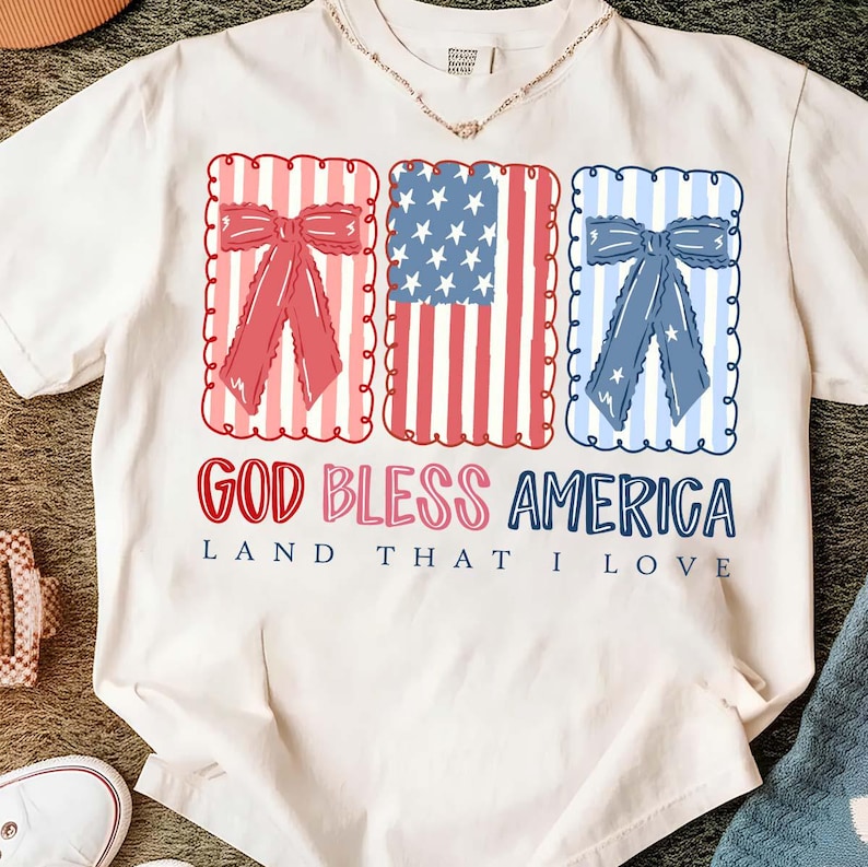 May include: White t-shirt with a patriotic design. The shirt features three rectangular frames with red, white, and blue elements, including bows and the American flag. The text "GOD BLESS AMERICA LAND THAT I LOVE" is printed below the frames.