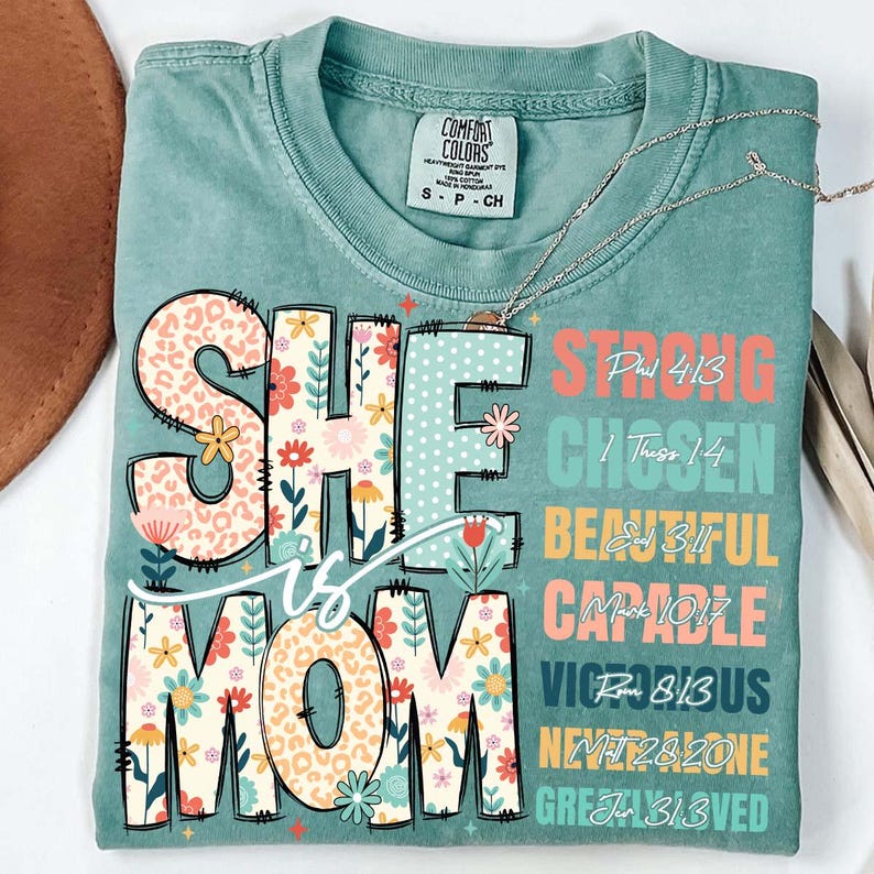 K&ouml;nnte beinhalten: Ein blaues T-Shirt mit einem floralen Design, das "She is Mom" sagt, mit Bibelversen um das Design herum. Die Verse sind: Phil 4:13 Strong, 1 Thess 1:4 Chosen,  Ezk 3:11 Beautiful, Mark 10:17 Capable, Rom 8:13 Victorious, Matt 28:20 Never Alone, Jn 3:13 Greatly Loved.