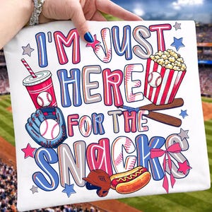 May include: White baseball-themed graphic featuring the text "I'M JUST HERE FOR THE SNACKS" in blue, pink, and white. The design includes a baseball, popcorn, hot dog, glove, drink, and baseball helmet.