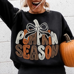 May include: A black long-sleeve sweatshirt featuring a graphic design of a pumpkin with a white bow on top. The text PUMPKIN SEASON is printed in a stylized, layered font using white, orange, and brown colors. The design is centered on the chest.