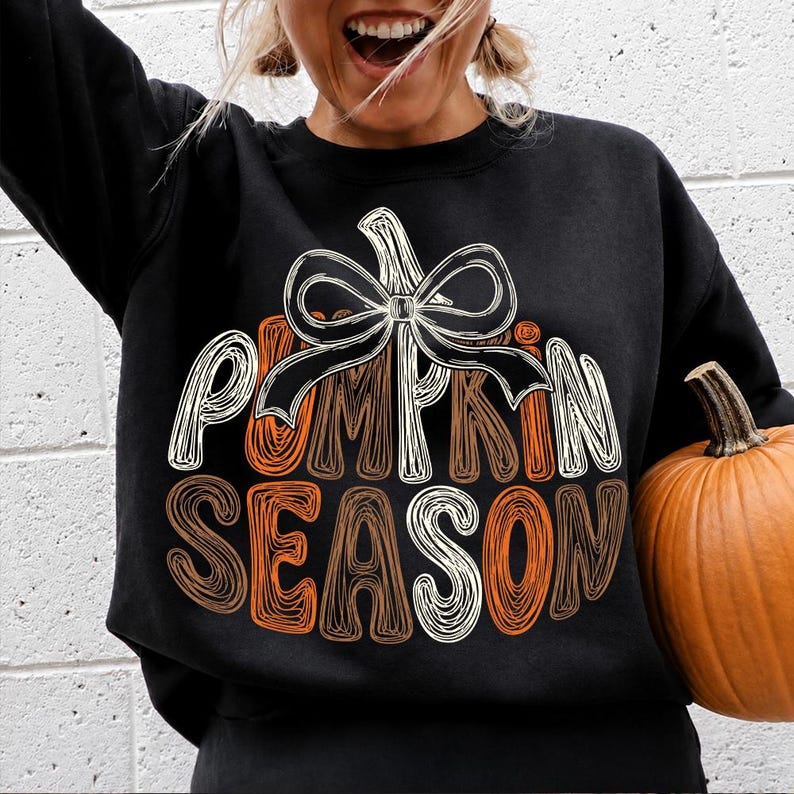 May include: A black long-sleeve sweatshirt featuring a graphic design of a pumpkin with a white bow on top. The text PUMPKIN SEASON is printed in a stylized, layered font using white, orange, and brown colors. The design is centered on the chest.