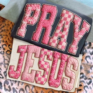 Puede incluir: Dos camisetas con texto bordado. La camiseta superior es azul marino con la palabra "PRAY" en rosa, algunas letras son a cuadros. La camiseta inferior es blanca con la palabra "JESUS" en rosa. Ambas camisetas tienen lazos blancos.