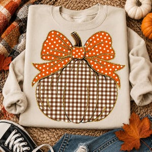 May include: A beige crewneck sweatshirt featuring a large graphic of a pumpkin with a brown and white gingham pattern. The pumpkin is topped with a bright orange bow decorated with white polka dots and outlined in a shimmering gold glitter effect.
