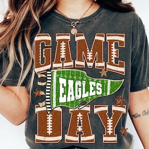 Puede incluir: Una camiseta gris carbón con el texto GAME DAY en letras grandes de color marrón con patrones de cordones de fútbol americano en blanco. Un banderín verde con la palabra EAGLES en blanco está centrado entre las palabras, con estrellas marrones.