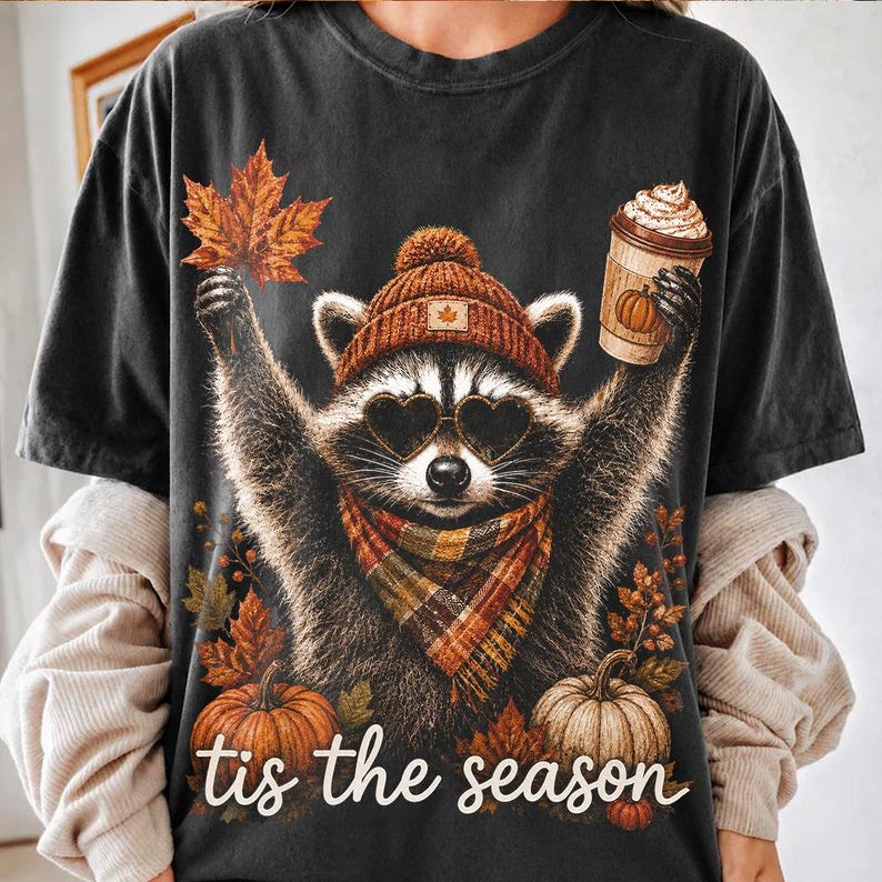 K&ouml;nnte beinhalten: Ein schwarzes T-Shirt mit einer Grafik eines Waschb&auml;ren, der eine braune Strickm&uuml;tze, herzf&ouml;rmige Sonnenbrillen und einen karierten Schal tr&auml;gt. Der Waschb&auml;r h&auml;lt ein Ahornblatt und einen Kaffeebecher. K&uuml;rbisse und Herbstbl&auml;tter umrahmen den Text "tis the season".