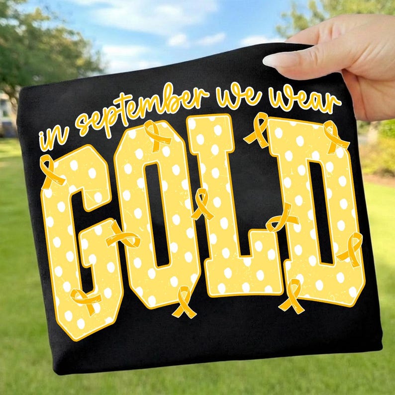 May include: A black t-shirt featuring the yellow text "in september we wear GOLD" in a varsity-style font. The letters are filled with a white polka dot pattern and decorated with small yellow awareness ribbons throughout the design.
