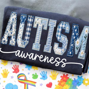 May include: Navy blue sweatshirt with the word "AUTISM" in patchwork letters, featuring floral, gingham, and polka dot patterns. The word "awareness" is written in white script below. Colorful handprints, puzzle pieces, and a rainbow ribbon are visible.