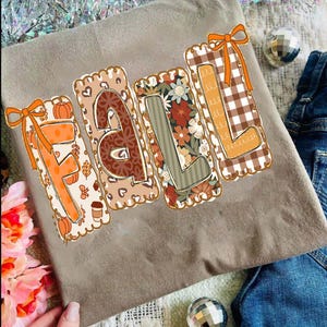 Peut inclure: Un t-shirt beige avec le mot "FALL" dans un design décoratif. Chaque lettre présente un motif différent, notamment des citrouilles, un imprimé léopard, des motifs floraux et du vichy. Les lettres ont un bord festonné et sont agrémentées de nœuds orange.