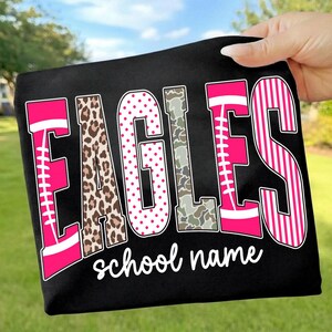 May include: A black t-shirt featuring the word EAGLES in large, multi-patterned letters. The letters include pink football designs, leopard print, pink polka dots, camouflage, and pink stripes. Below the word, the text school name is printed in white.
