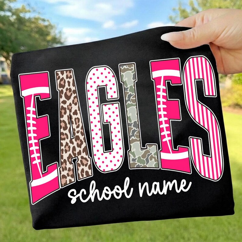 May include: A black t-shirt featuring the word EAGLES in large, multi-patterned letters. The letters include pink football designs, leopard print, pink polka dots, camouflage, and pink stripes. Below the word, the text school name is printed in white.