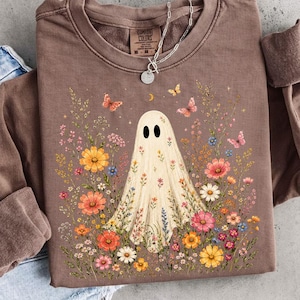May include: A brown long-sleeve crewneck sweatshirt featuring a central graphic of a white ghost surrounded by colorful wildflowers and butterflies. The garment has a ribbed collar with a tag reading Comfort Colors and a silver chain necklace.