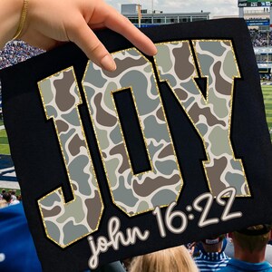 May include: Black sign with the word "JOY" in large, gold-outlined letters filled with a camouflage pattern of gray, brown, and green. The text "john 16:22" is written in a cursive font below the word "JOY".