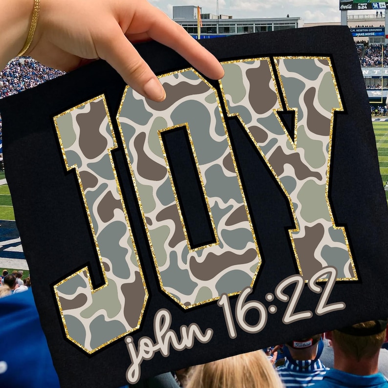 May include: Black sign with the word "JOY" in large, gold-outlined letters filled with a camouflage pattern of gray, brown, and green. The text "john 16:22" is written in a cursive font below the word "JOY".