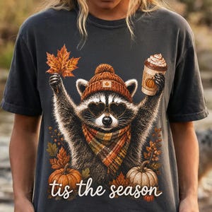 K&ouml;nnte beinhalten: Ein anthrazitfarbenes T-Shirt mit einer Grafik eines Waschb&auml;ren, der eine braune Strickm&uuml;tze, herzf&ouml;rmige Sonnenbrillen und einen karierten Schal tr&auml;gt. Der Waschb&auml;r h&auml;lt ein Ahornblatt und einen Kaffeebecher. K&uuml;rbisse und Bl&auml;tter umgeben den Text 'tis the season'.