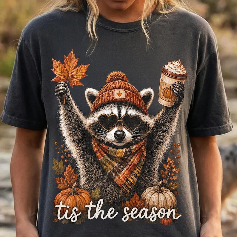 K&ouml;nnte beinhalten: Ein anthrazitfarbenes T-Shirt mit einer Grafik eines Waschb&auml;ren, der eine braune Strickm&uuml;tze, herzf&ouml;rmige Sonnenbrillen und einen karierten Schal tr&auml;gt. Der Waschb&auml;r h&auml;lt ein Ahornblatt und einen Kaffeebecher. K&uuml;rbisse und Bl&auml;tter umgeben den Text 'tis the season'.