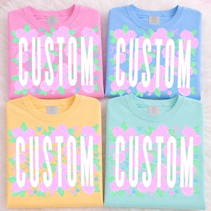 May include: Four t-shirts in pink, blue, yellow, and teal, each with a floral design and the word "CUSTOM" in white. The floral design features pink flowers and green leaves. The shirts are neatly folded.