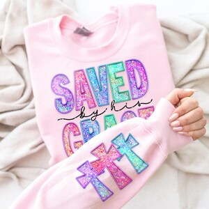 May include: A light pink sweatshirt with the words "SAVED BY HIS GRACE" in colorful glitter lettering. The sleeve features three glitter cross designs in shades of pink, purple, and blue.