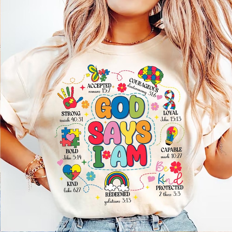 May include: A white t-shirt with a colorful graphic design that says "God Says I Am." The graphic includes a rainbow, a heart, a lightbulb, a butterfly, and a handprint. The words "Strong, Bold, Kind, Accepted, Courageous, Loyal, Capable, Be Kind, Protected, Redeemed" are also included in the graphic, along with corresponding Bible verses.