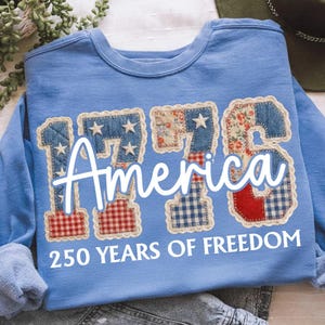 Pode incluir: Camisola azul clara com o texto "1776 America" em escrita branca. Os números são patchwork com padrões de ganga, florais e xadrez. O texto "250 YEARS OF FREEDOM" está por baixo.