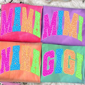 May include: Four colorful t-shirts with the names "MIMI", "NANA", and "GIGI" in bold, leopard print lettering. The shirts are in shades of pink, purple, orange, and teal. The letters are outlined in bright colors. The shirts are on a white fluffy surface.