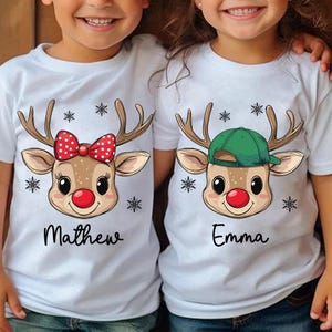 May include: Two white t-shirts featuring a reindeer design. One shirt has a red bow on the reindeer and says "Matthew". The other shirt has a green hat on the reindeer and says "Emma".
