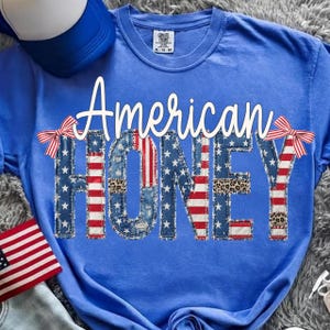 Può includere: Maglietta blu con la scritta "American HONEY" in corsivo bianco. Le lettere sono decorate con stelle, strisce e stampa leopardata, con fiocchi rossi e bianchi su entrambi i lati.