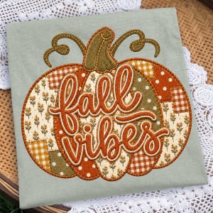 May include: A sage green t-shirt featuring a large, central pumpkin applique. The pumpkin is constructed from various fabric patches in orange, white, and green with floral, gingham, and polka dot patterns. The text fall vibes is embroidered.