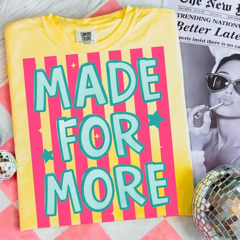 May include: Yellow t-shirt with the text "MADE FOR MORE" in turquoise letters, set against pink vertical stripes. The shirt has a crew neck and a small Comfort Colors logo.