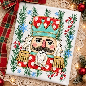 May include: A white fabric square featuring a central illustration of a nutcracker wearing a red and white polka dot hat and jacket with a teal crown. The design includes green pine branches, blue snowflakes, and red and white candy canes.