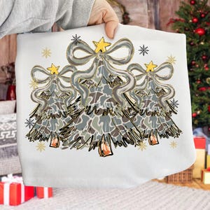 Può includere: Borsa tote bianca con tre alberi di Natale stilizzati con motivi grigi, marroni e verdi. Ogni albero ha un fiocco argentato, una stella dorata e dettagli di fiocchi di neve dorati. La borsa è tenuta da una persona.