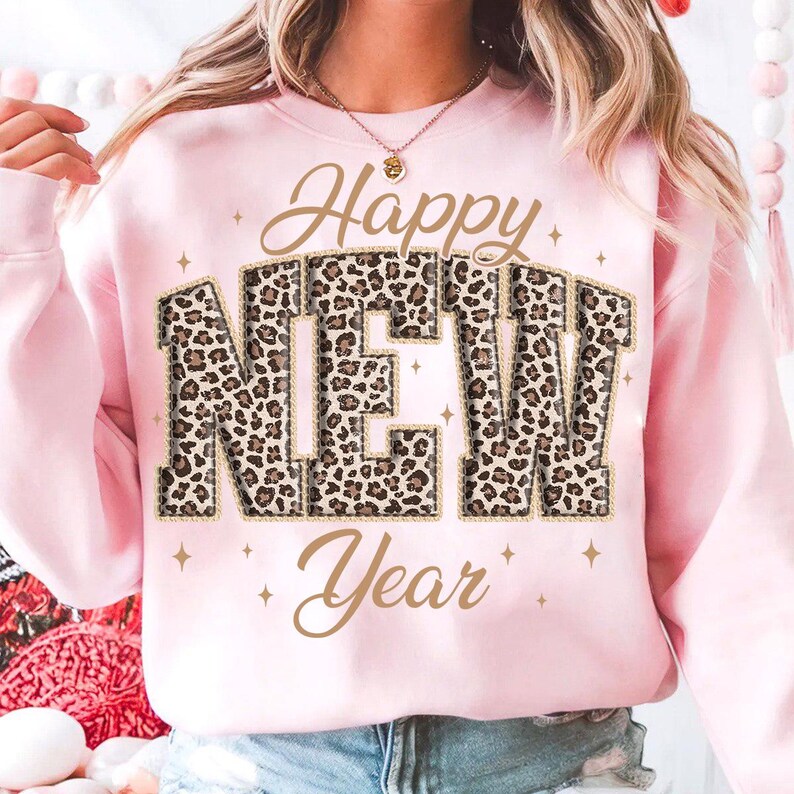 K&ouml;nnte beinhalten: Ein hellrosa Sweatshirt mit den Worten "Happy NEW Year" in goldener Schrift und Leopardenmuster. Das Sweatshirt hat einen Rundhalsausschnitt und lange &Auml;rmel. Kleine goldene Sterne sind um den Text herum verstreut.
