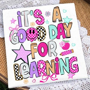 May include: A white square fabric panel featuring the colorful text IT'S A GOOD DAY FOR LEARNING. The letters are decorated with various patterns including checkers, stripes, and polka dots in pink, purple, teal, and tan, with a smiley face and pencil.