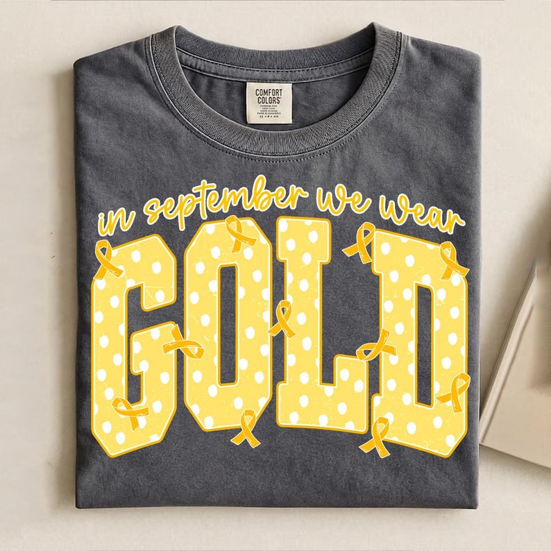 May include: A charcoal gray crew neck t-shirt featuring the yellow text "in september we wear GOLD" in a collegiate font. The letters are filled with a white polka dot pattern and decorated with small yellow awareness ribbons throughout.