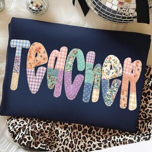 Pode incluir: Camisola azul marinho com a palavra "TEACHER" aplicada em tecido patchwork colorido. As letras apresentam padrões florais, de bolinhas e xadrez. Um tecido com estampa de leopardo é visível na parte inferior.