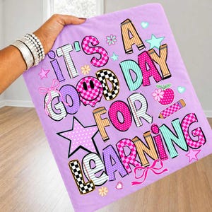May include: A light purple fabric square featuring the colorful text IT'S A GOOD DAY FOR LEARNING. The letters are decorated with various patterns including checkers, stripes, and polka dots, alongside graphic elements like stars, bows, and a pencil.