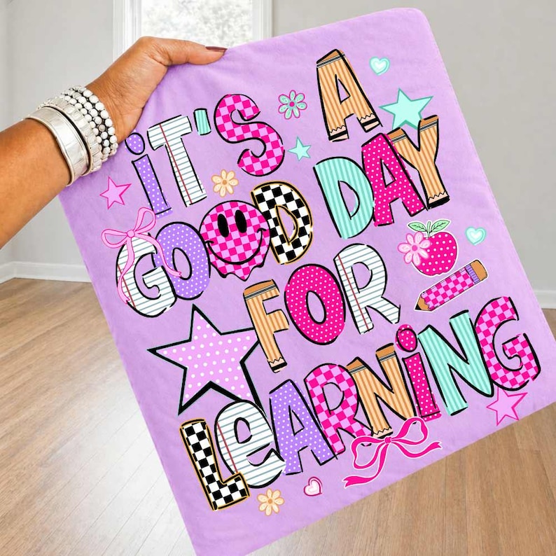 May include: A light purple fabric square featuring the colorful text IT'S A GOOD DAY FOR LEARNING. The letters are decorated with various patterns including checkers, stripes, and polka dots, alongside graphic elements like stars, bows, and a pencil.