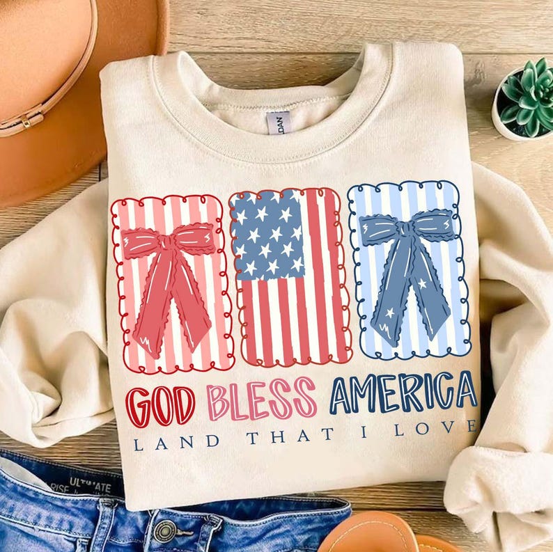 May include: Cream-colored sweatshirt with a patriotic design. Features three rectangular graphics: a red bow, an American flag, and a blue bow. The text "GOD BLESS AMERICA LAND THAT I LOVE" is printed below the graphics.