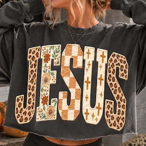 May include: A charcoal gray long-sleeve cropped sweatshirt featuring the word JESUS in large block letters. Each letter has a unique pattern: leopard print, floral print with orange flowers, tan and white checkerboard, and gold crosses.