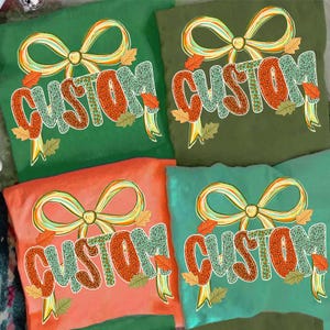 May include: Four colorful t-shirts with the word "CUSTOM" in a fall-themed design. Each shirt features a large bow and autumn leaves. The shirts are in shades of green, orange, and teal.