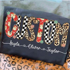 May include: Dark gray t-shirt with the word "CUSTOM" in a festive design. The letters are decorated with Santa Claus, leopard print, and a black and white checkerboard pattern. Names are printed below the word "CUSTOM".