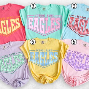 May include: Six short-sleeved crew-neck t-shirts in pink, mint, light blue, lavender, yellow, and light pink. Each shirt features the text SCHOOL NAME above the word EAGLES in a block font with a gingham pattern, enclosed in a geometric frame.