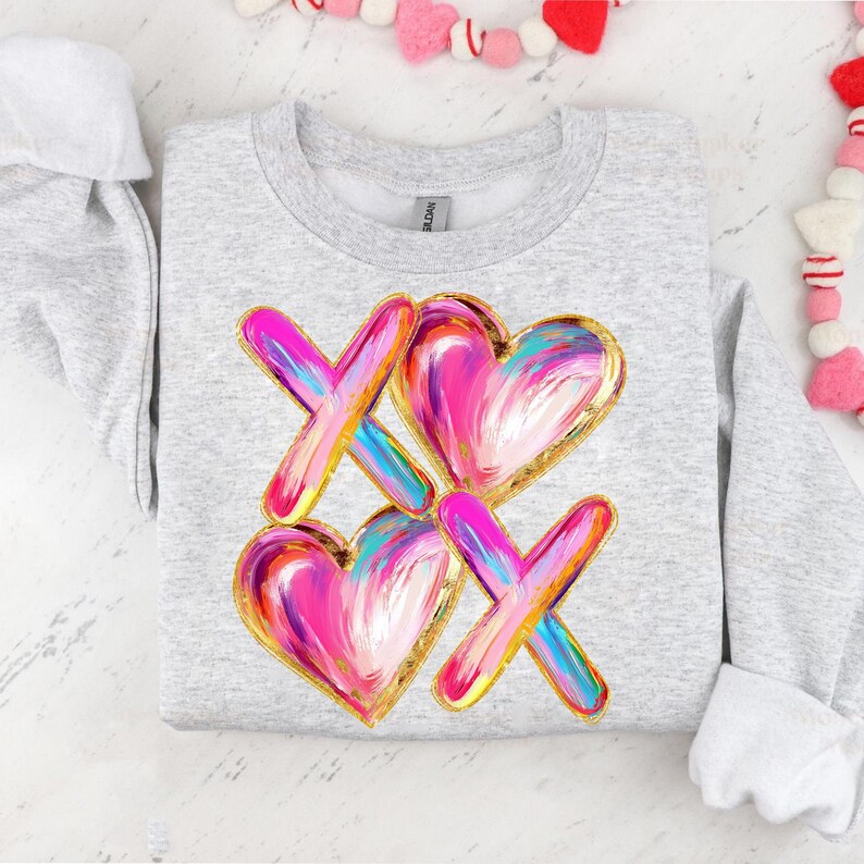 Puede incluir: Una sudadera gris con un dise&ntilde;o rosa y dorado de corazones y equis. El dise&ntilde;o est&aacute; pintado en un estilo de pinceladas.