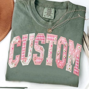 May include: An olive green t-shirt with the word "CUSTOM" appliqued in pink and white fabric. The letters feature floral, gingham, and polka dot patterns. A gold necklace with a pendant rests on the shirt.