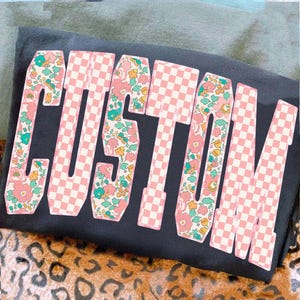 May include: A black t-shirt featuring the word "CUSTOM" in large, eye-catching letters. The letters alternate between a pink and white chequered pattern and a floral design with pink, yellow, and teal flowers. The shirt is folded.