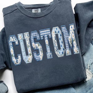 May include: A blue sweatshirt with the word "CUSTOM" appliqued across the front. The letters are a patchwork of floral, gingham, and polka dot patterns in shades of blue and white. The tag reads "COMFORT COLORS".