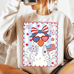 Puede incluir: Una sudadera blanca con un dise&ntilde;o de ganso patri&oacute;tico. El ganso lleva gafas de sol en forma de coraz&oacute;n, un lazo y una diadema rojos, blancos y azules. Sostiene una bandera estadounidense, con fuegos artificiales y estrellas de fondo.