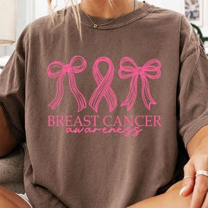 May include: A brown short-sleeve t-shirt featuring a bright pink graphic design. The design includes two bows flanking a central awareness ribbon, with the text BREAST CANCER awareness printed in a combination of block and script lettering.