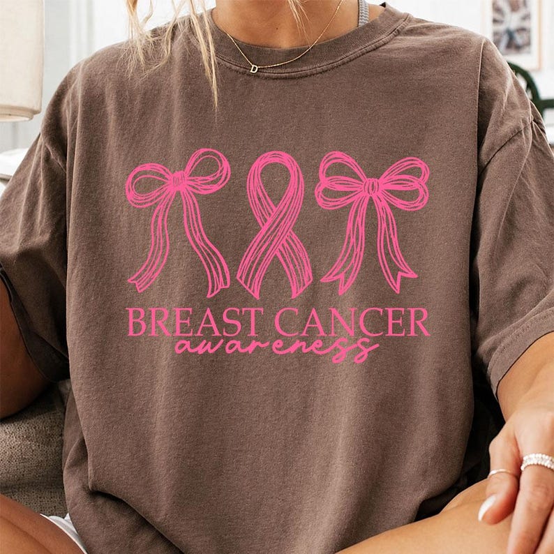 May include: A brown short-sleeve t-shirt featuring a bright pink graphic design. The design includes two bows flanking a central awareness ribbon, with the text BREAST CANCER awareness printed in a combination of block and script lettering.