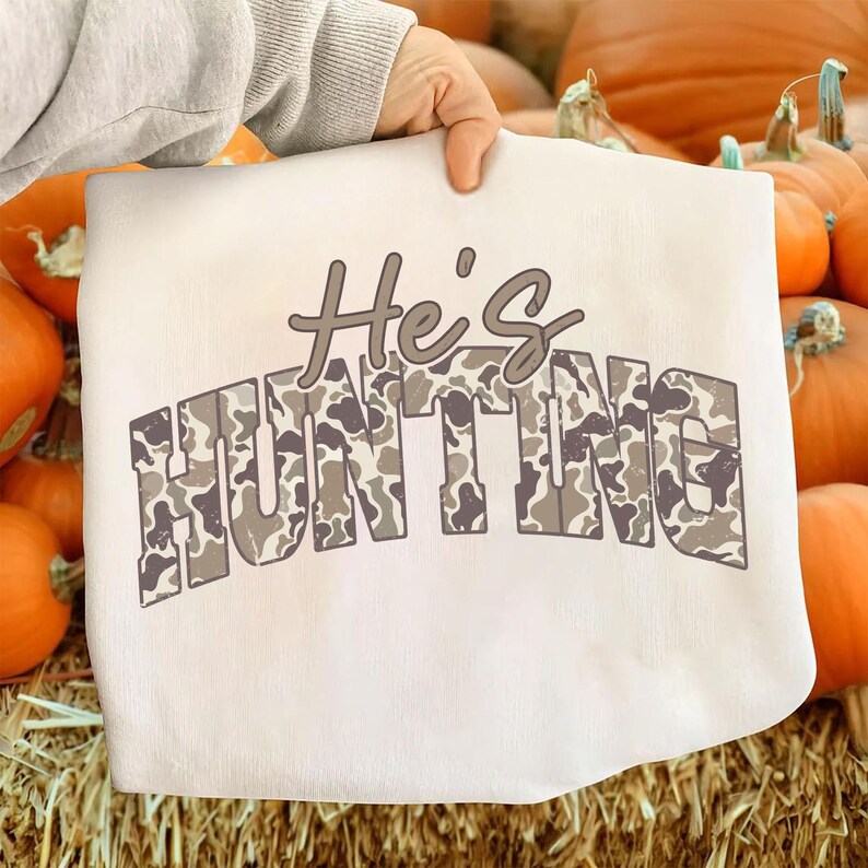 May include: A white sweatshirt with the text "He's HUNTING" in a camouflage pattern. The sweatshirt is held up in front of a background of pumpkins.