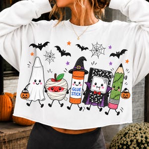 Pode incluir: Uma camisola branca de manga comprida com um gr&aacute;fico colorido de cinco personagens de desenhos animados: um fantasma, uma ma&ccedil;&atilde; m&uacute;mia, uma cola em barra com chap&eacute;u de bruxa, um caderno vampiro e um l&aacute;pis, rodeados por morcegos e estrelas.